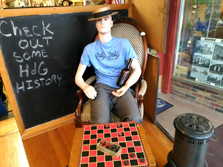 Bahoukas front window display with man on chair, playing checkers, nice heater to the side, blackboard behind him, warm and cozy in January